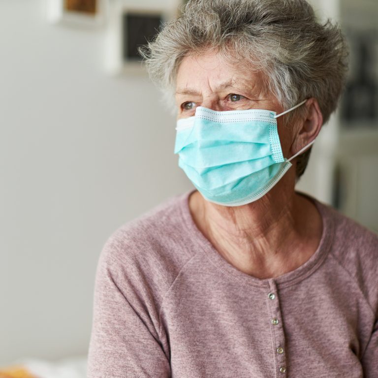 A senior citizen sits alone on her bed with a respirator or surgical mask and looks sadly and frightened out of the window and into the camera
